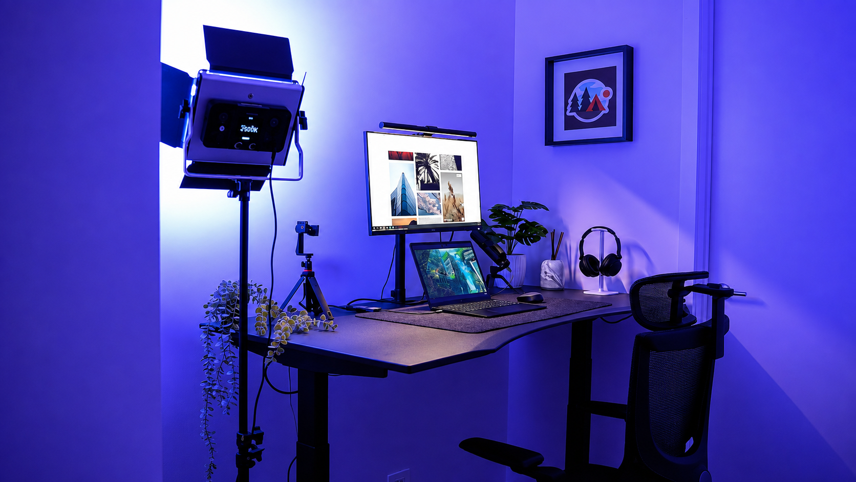 A modern standing desk setup in a home office environment, illuminated by blue ambient lighting. The desk is equipped with a dual-monitor setup (one desktop monitor and one laptop), a black office chair, a studio softbox light on a stand, and small potted plants.